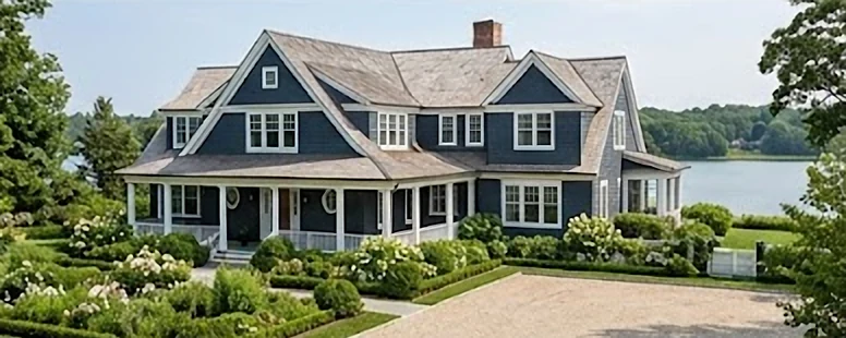 A navy shingle house with a wraparound porch and gardens, with water beyond.